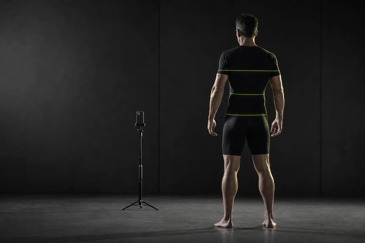 Un hombre de espaldas, con ropa deportiva ceñida, de pie frente a un móvil colocado en un trípode; sobre su cuerpo se proyectan líneas de medición verdes a la altura del pecho, la cintura y la cadera.