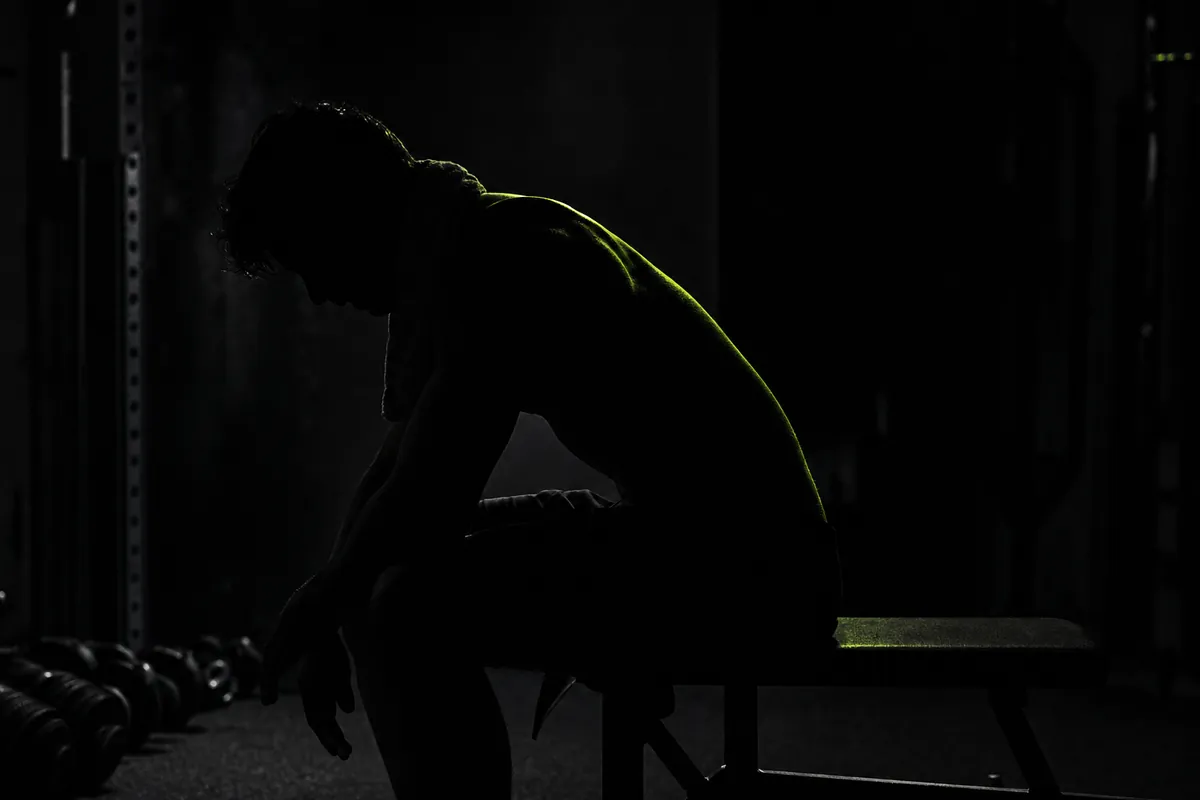 Un hombre sentado en un banco de gimnasio en penumbra, recuperando el aliento con una toalla al cuello, a contraluz.