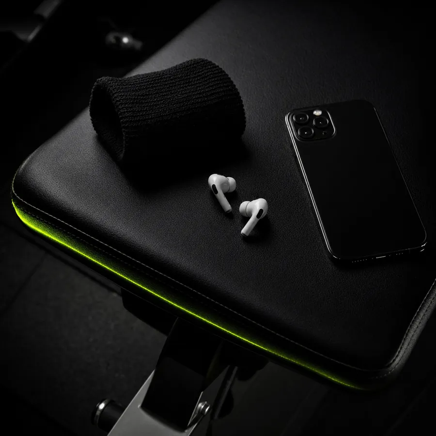 Unos auriculares inalámbricos y un teléfono móvil sobre un banco de gimnasio negro, con un filo de luz verde lima en el canto.